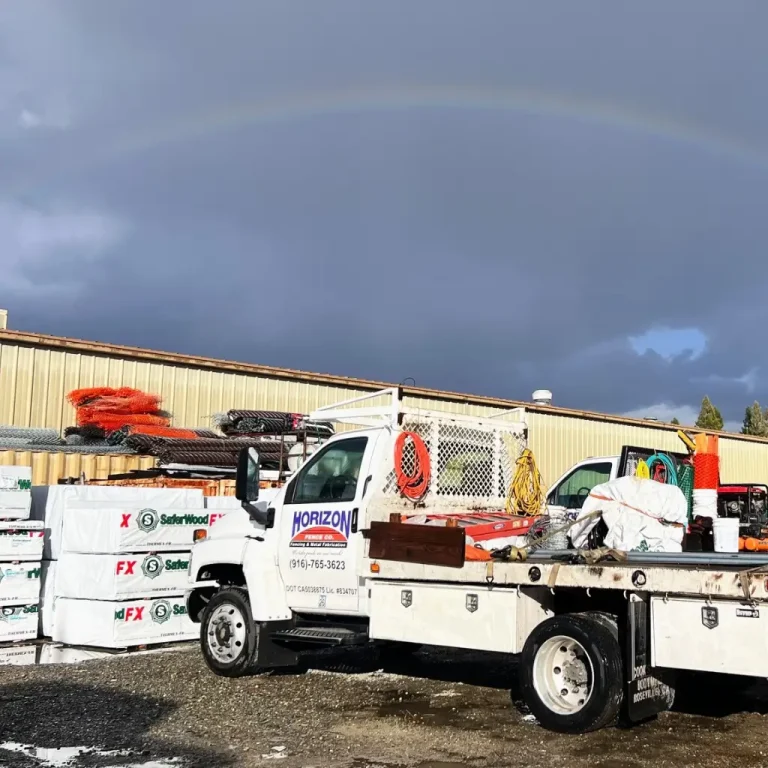 A Horizon Fence Company work truck loaded with tools and materials parked in the company yard, with fencing supplies stacked nearby and a faint rainbow in the cloudy sky above.