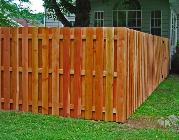 Redwood shadowbox fence with alternating vertical panels allowing airflow and filtered light along a property line with trees.