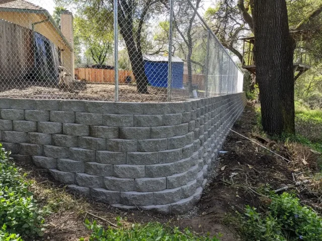 A chain link fence installed atop a retaining wall in a residential Orangevale backyard, enclosing a safe area for dogs with trees and a wooden structure nearby.