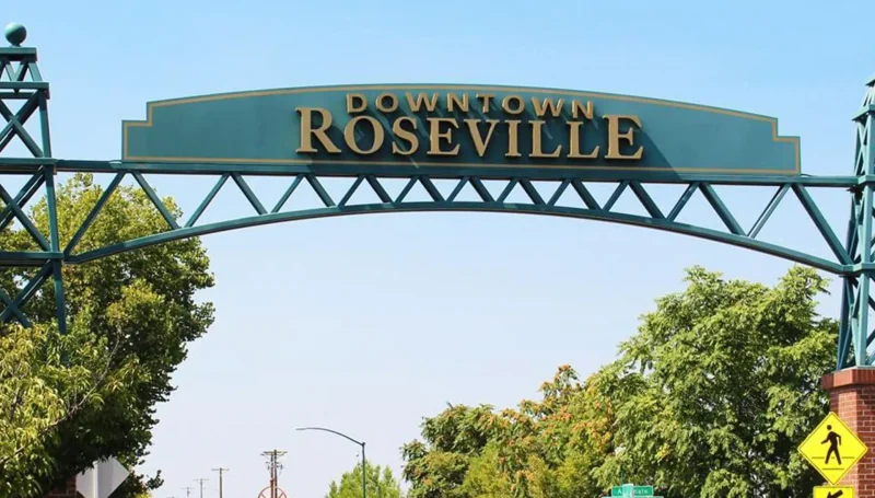 A wide-angle shot of the Downtown Roseville archway sign, painted teal with gold letters, over a street with a clear blue sky and lush green trees.