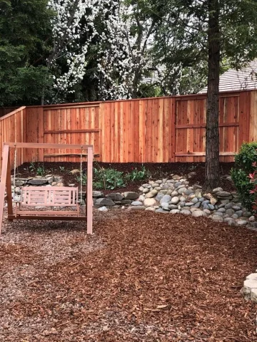 Cedar wood privacy fence installed in a landscaped Orangevale backyard featuring a wooden swing, river rock border, and blooming trees.